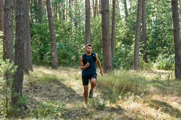Young caucasian sportsman running or jogging in green forest. Handsome bearded dark-haired athletic man wearing sportswear. Concept of modern healthy lifestyle. Sunny summer daytime
