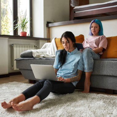 Kız kanepede dijital tablet izlerken kız arkadaşı evde dizüstü bilgisayar kullanıyor ve daktilo yazıyor. Kafkasyalı genç lezbiyen çift. Ev hayatı, eğlence ve boş zaman. Modern dairede oturma odası