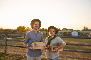 Köydeki ya da çiftlikteki otlakların yanında organik mısırlı ahşap sepetler tutan genç, beyaz çiftçi çifti. Taze hasat. Modern çiftlik hayatı. Tarım. Kırsal alan. Gün batımı zamanı