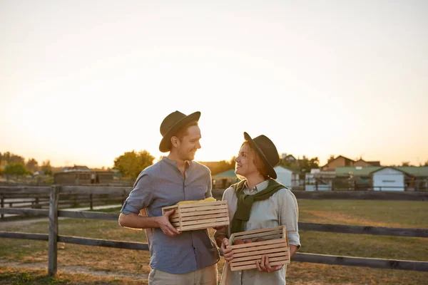 Young smiling caucasian farmer couple holding wooden baskets with organic corn and carrot and looking at each other in village or ranch. Fresh harvest. Modern farm lifestyle. Agriculture. Sunset time