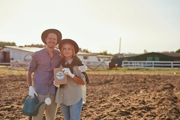 Young smiling caucasian farmer couple holding watering can and seeds on plowing field on ranch or farm. Eco harvest. Environment sustainability. Modern countryside lifestyle. Agriculture. Sunny day