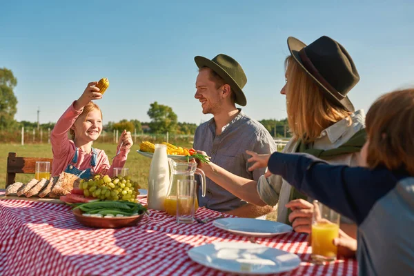 Young happy caucasian hipster farm family having lunch with fresh organic products in countryside outdoors. Mother giving plate with vegetables for daughter with corn. Modern farm lifestyle. Sunny day