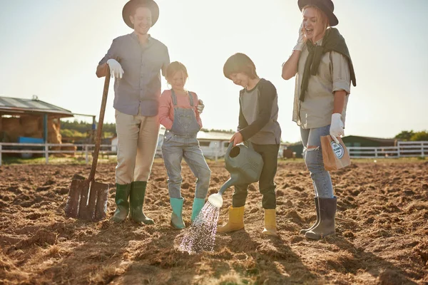 Mother, father and daughter looking at son watering seeds in ground hole on plow field on ranch or farm. Young caucasian hipster family. Eco harvest. Modern village lifestyle. Agriculture. Sunny day