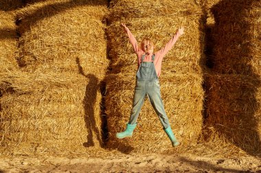 Çiftlikte saman balyalarıyla ahırda zıplayan elleri havada neşeli beyaz bir kız. Kameraya bakan bir kadın çocuk. Modern kırsal yaşam tarzı. Tarım. Sıcak güneşli bir gün.