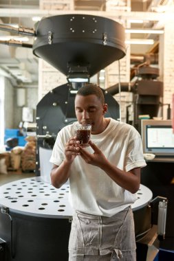 Profesyonel erkek barista fabrikada bardakta taze organik kahve kokluyor. Genç, odaklanmış, gözleri kapalı bir siyah adam kahve aromasını kontrol ediyor ve inceliyor. Kahve yapımı. Küçük işletme