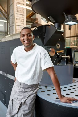 Fabrikadaki endüstriyel kahve çekirdeği kızartma makinesinin yanında gülümseyen siyah bir erkek işçi. Kahve ve üretim. Küçük bir iş. Modern otomatik üretim ekipmanları. Adam kameraya bakıyor.