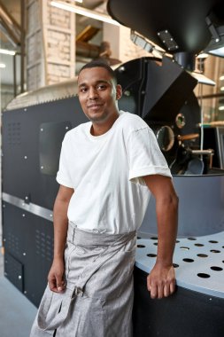 Male barista or worker near industrial coffee bean roasting machine on factory. Coffee making and production. Small business. Modern automated manufacturing equipment. Black man looking at camera