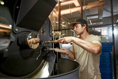 Male barista or worker pouring out coffee beans in roasting machine during work on factory. Coffee process making and production. Small business. Modern manufacturing equipment. Young caucasian man