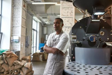 Genç siyah erkek işçi ya da fabrikadaki endüstriyel kahve çekirdeği kızartma makinesinin yanındaki işyeri sahibi. Kahve yapımı ve üretimi. Küçük bir iş. Modern otomatik üretim ekipmanları