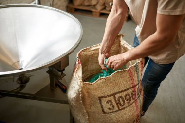 Partial male barista or worker opening package in sack with coffee beans for pouring out in metal grinder. Coffee process making and production. Small business. Modern manufacturing equipment