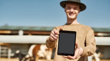 Dijital tabletin seçmeli odak noktası çiftlikte arka planda bulanık süt inekleri olan erkek çiftçinin ellerinde. Genç beyaz bir adam kameraya bakıyor. Modern kırsal yaşam tarzı. Tarım