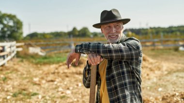 Çiftlikte ya da çiftlikte çalışırken tarımsal aletleri olan gri saçlı bir erkek çiftçi dinleniyor ve başka tarafa bakıyor. Kovboy şapkası takan odaklanmış beyaz bir adam. Modern kırsal yaşam tarzı. Tarım. Güneşli bir gün