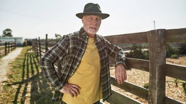 Çiftlikteki ya da çiftlikteki çitlere bakan yaşlı bir erkek çiftçi. Odaklanmış, elleri kalçasında, şapkalı, beyaz bir adam. Modern kırsal yaşam tarzı kavramı. Tarım ve tarım. Sıcak güneşli bir gün.