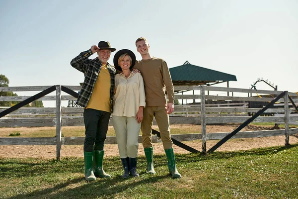 Middle aged farmer couple and their teenage grandson hug and look at camera on farm or ranch. Smiling caucasian family enjoying time together. Modern countryside lifestyle. Agriculture. Warm sunny day