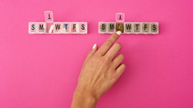 Partial of woman hand taking dry marijuana bud from smtwtfs package near pills. Light drug and addiction. Herbal medicine and painkiller therapy. Natural organic cannabis. Pink background. Copy space
