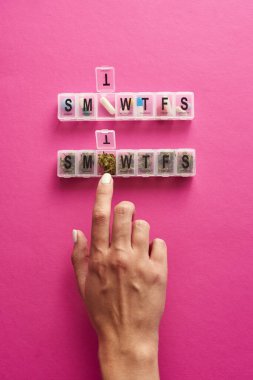 Cropped of female hand taking dry marijuana bud from smtwtfs package near pills. Light drug and addiction. Herbal medicine and painkiller therapy. Natural homemade organic cannabis. Pink background
