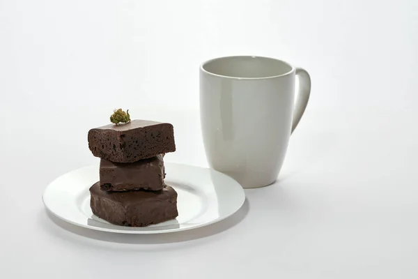 Cup with coffee and green dry crushed marijuana bud on stack of sweet tasty chocolate cake pieces on plate. Light drug and addiction. Natural organic cannabis. Isolated on white background. Copy space