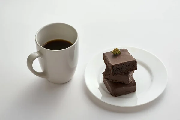Cup with coffee and fresh green dry crushed marijuana bud on sweet chocolate cake pieces on plate. Light drug and addiction. Natural organic cannabis. Isolated on white background. Copy space