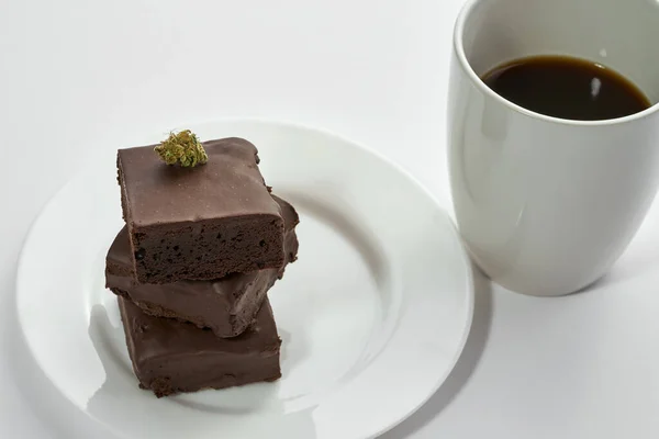 Cropped cup with coffee and fresh green dry crushed marijuana bud on stack of sweet chocolate cake pieces on plate. Light drug and addiction. Natural organic cannabis. Isolated on white background