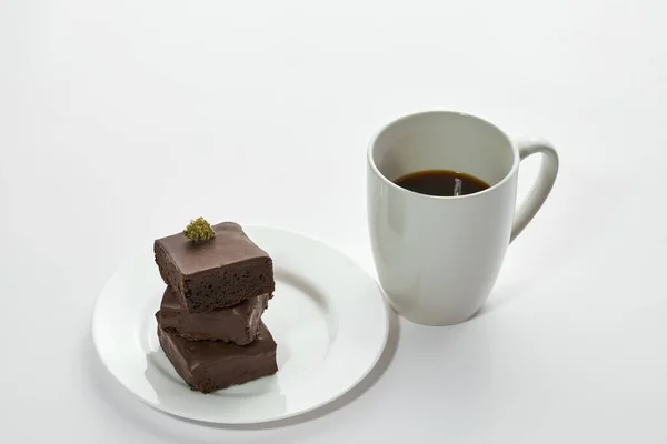 Cup with coffee and fresh green dry crushed marijuana bud on stack of sweet chocolate cake pieces on plate. Light drug and addiction. Natural organic cannabis. Isolated on white background. Copy space