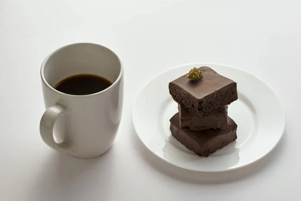 Cup with coffee and green dry crushed marijuana bud on stack of sweet chocolate cake pieces on plate. Light drug and addiction. Natural organic cannabis. Isolated on white background. Copy space