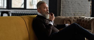 Geniş dairedeki kanepede tütün piposu tüttüren son derece düşünceli beyaz bir adam. Sıradan kıyafetler giyen şık sakallı bir emekli. Modern yaşlı erkek yaşam tarzı. Ev istirahatı ve boş zaman