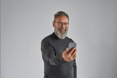 Akıllı telefondan video görüşmesi yapan gülümseyen yaşlı Avrupalı bir adam. Sakallı emekli süveter, gözlük ve kablosuz kulaklık takıyor. Modern son sınıf erkek yaşam tarzı. Gri arka plan. Stüdyo çekimi. Boşluğu kopyala