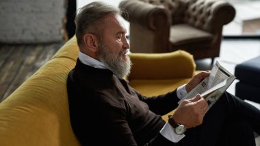 Geniş bir dairede, koltukta günlük okuyan gri saçlı, beyaz bir adamın yan görüntüsü. Sıradan kıyafetler giyen modaya uygun sakallı bir emekli. Modern son sınıf erkek yaşam tarzı. Ev içi istirahat ve rahatlık.