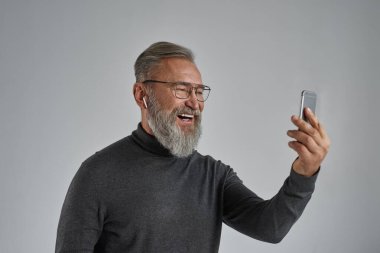 Kıdemli gülümseyen beyaz adam akıllı telefondan izliyor. Süveter, gözlük ve kablosuz kulaklık takan şık sakallı emekli. Modern yaşlı erkek yaşam tarzı. Gri arka plan. Stüdyo çekimi. Boşluğu kopyala