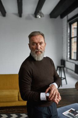 Yaşlı, ciddi, beyaz bir adam kol saatini düzeltiyor ve geniş bir dairede kameraya bakıyor. Sıradan kıyafetler giyen modaya uygun sakallı bir emekli. Modern son sınıf erkek yaşam tarzı kavramı. Gündüz