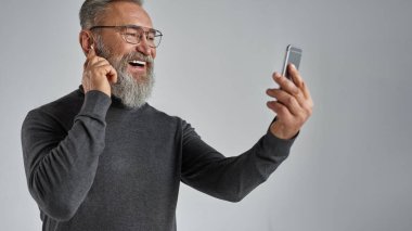 Akıllı telefondan müzik seçmek için kablosuz kulaklık kullanan Avrupalı yaşlı bir adam. Süveter ve gözlük takan emekli. Modern son sınıf erkek yaşam tarzı. Gri arka plan. Stüdyo çekimi. Boşluğu kopyala
