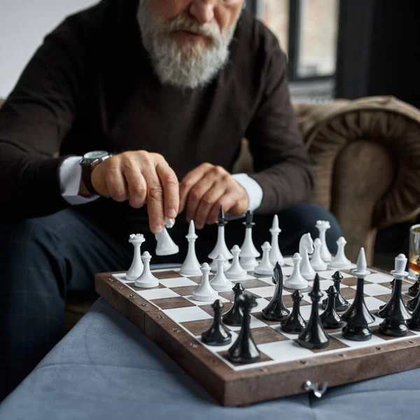 Obscure face of senior man playing chess with himself in armchair in spacious apartment. Stylish bearded pensioner. Modern senior male lifestyle. Domestic entertainment, hobby and leisure