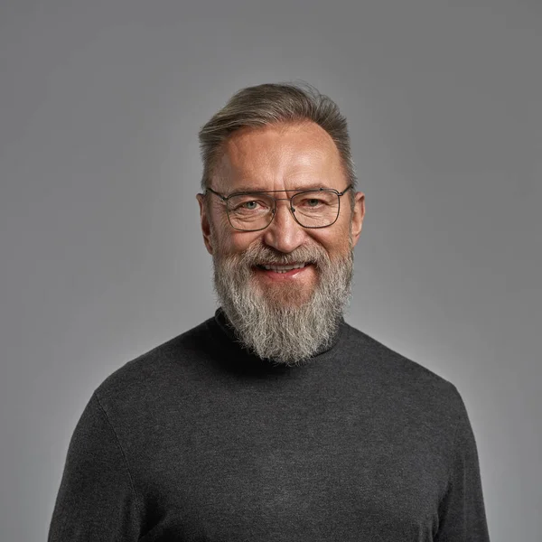 Portrait of grey hair smiling caucasian man looking at camera. Fashionable bearded pensioner wearing glasses and sweater. Modern elderly male lifestyle. Grey background. Studio shoot. Copy space