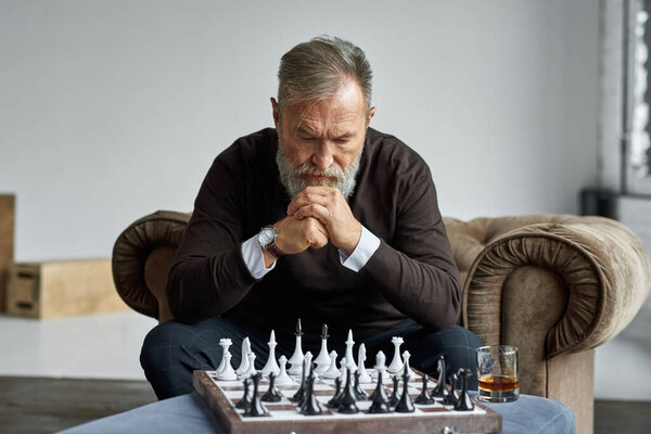 Senior thoughtful caucasian man playing chess with himself in armchair in spacious apartment. Stylish bearded pensioner. Modern senior male lifestyle. Domestic entertainment, hobby and leisure