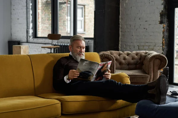 Elderly focused caucasian man reading journal on sofa at home. Trendy bearded pensioner wearing casual clothes. Modern senior male lifestyle. Domestic rest and leisure. Spacious apertment interior