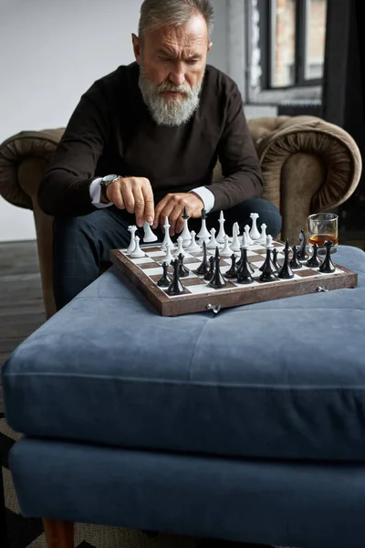 Grey hair thoughtful caucasian man playing chess with himself in armchair at home. Fashionable bearded pensioner. Modern senior male lifestyle. Domestic entertainment, hobby and leisure. Spacious flat