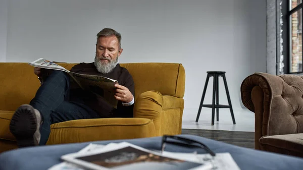 Elderly Concentrated Caucasian Man Reading Newspaper Sofa Spacious Apartment Trendy — Foto Stock