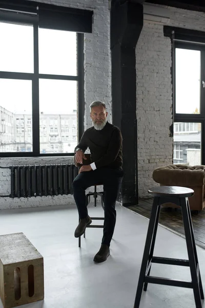 Elderly confident caucasian man sitting on high chair and looking at camera in spacious apartment. Trendy bearded pensioner wearing casual clothes. Concept of modern senior male lifestyle. Daytime