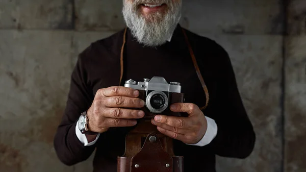 Obscure face of senior smiling man with old camera. Stylish bearded pensioner. Modern elderly male lifestyle. Entertainment and hobby. Isolated on rusty concrete background. Studio shoot