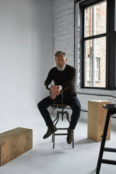 Senior confident caucasian man sitting on high chair and looking at camera in spacious apartment. Stylish bearded pensioner wearing casual clothes. Concept of modern elderly male lifestyle. Daytime
