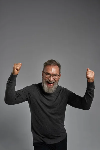 Excited grey hair caucasian man celebrating his success or win. Pleased bearded pensioner wearing glasses looking at camera. Modern elderly male lifestyle. Grey background. Studio shoot. Copy space