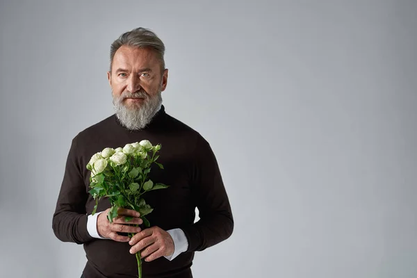 Elderly serious caucasian man with bouquet of white roses. Bearded male wearing sweater. Present for romantic meeting, Vaentines Day and 8 march. Isolated on grey background. Studio shoot. Copy space