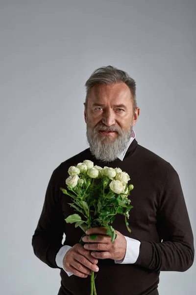 Grey hair serious caucasian man with bouquet of white roses. Fashionable bearded pensioner. Present for romantic meeting, Vaentines Day and 8 march. Isolated on grey background in studio. Copy space