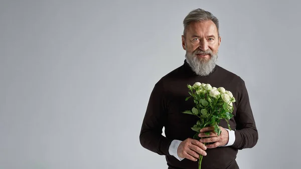 Senior european man with bouquet of white roses looking at camera. Stylish bearded male. Present for romantic meeting, Vaentines Day and 8 march. Isolated on grey background. Studio shoot. Copy space