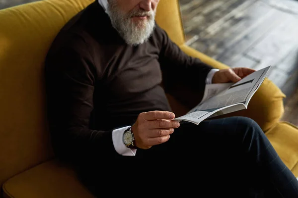 Obscure face of senior man reading journal on sofa in spacious apartment. Stylish bearded pensioner wearing wristwatch and casual clothes. Modern elderly male lifestyle. Domestic rest and leisure