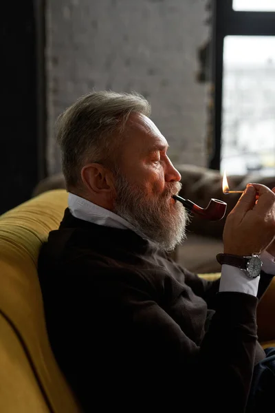 Senior Focused Caucasian Man Lighting Tobacco Pipe Match Sofa Spacious — Stockfoto
