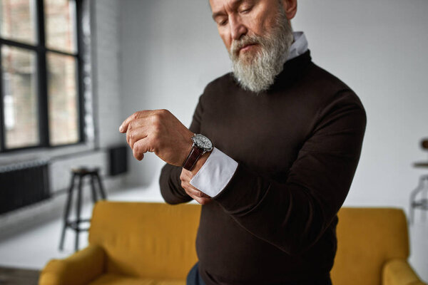 Cropped image of grey hair serious caucasian man correcting shirt sleeve in spacious flat. Fashionable bearded pensioner wearing casual clothes. Concept of modern elderly male lifestyle. Daytime