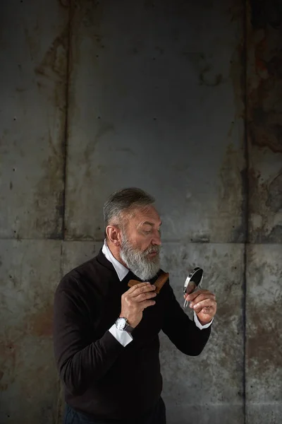 Stylish senior caucasian man looking at mirror of combing his beard with comb. Modern elderly male lifestyle. Personal beauty and hygiene. Isolated on rusty concrete background in studio. Copy space