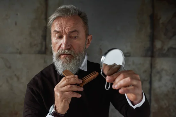 Elderly focused elegant caucasian man looking at camera of combing his beard with comb. Modern senior male lifestyle. Personal beauty and hygiene. Isolated on rusty concrete background. Studio shoot