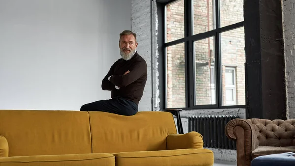 Grey hair focused caucasian man with crossed arms sitting on sofa top and looking away in spacious flat. Fashionable bearded pensioner wearing casual clothes. Concept of modern elderly male lifestyle